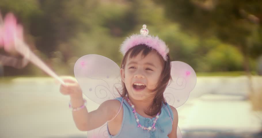 Cheerful little Asian girl in pink fairy queen costume playing make-believe in park in summer