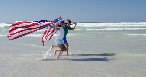 Siblings holding American flag while running on shore at beach on a sunny day 스톡 비디오