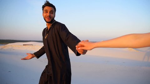 Handsome male Arab holding woman's hand, chatting and smiling looking at her. Lovers walk through expanses of sandy desert at sunset under open blue sky on summer evening. dark man with short dark