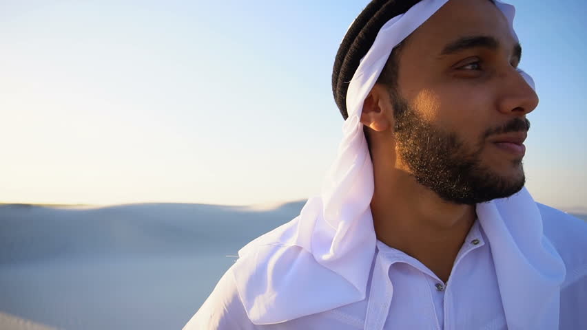 Close-up portrait of handsome Muslim young man who smiles and looks around, watching beauty of nature in sandy desert with white sand against blue sky in national suit that develops in wind. Swarthy Royalty-Free Stock Footage #29765752