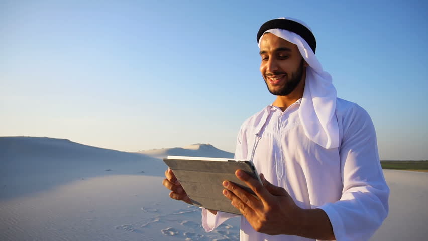 Portrait of stately Arabian Sheikh man who with smile on face holds tablet in hands. guy looks around desert and keeps record in gadget or writes down impressions of journey, standing in desert in