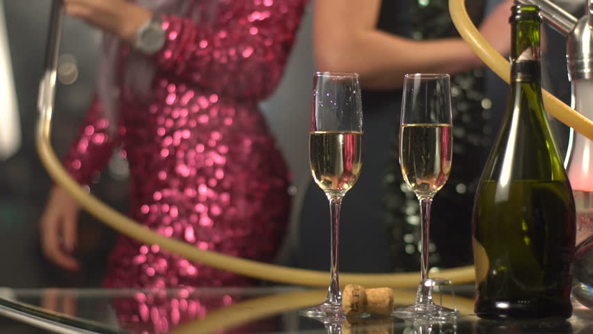 The composition consisting of two glasses with champagne and bottle placed on the table at the blurred background of two girlfriends smoking the hookah while dancing.