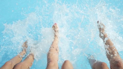 SLOW MOTION CLOSE UP: Three unrecognizable cheerful girls sitting on pool edge, splashing water with their feet on hot sunny day. Three playful young women on summer vacation splashing water with legs