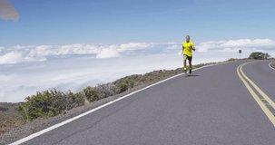 Fit male athlete running in slow motion on mountain road above the clouds. Determined young man is exercising in nature during sunny day.