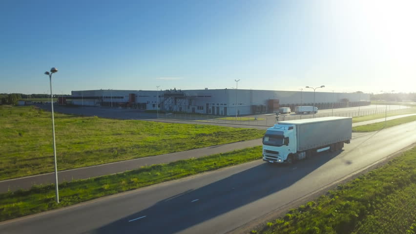 Aerial Follow Shot of White Truck with Semi Trailer Attached Moving Through Industrial Warehouse, Rural Area. Sunset. Shot on Phantom 4K UHD Camera.