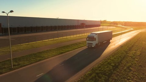 Aerial View of White Semi Truck with Cargo Trailer Moving on the Highway. In the Background Warehouses and Rural Area, Sun is Setting. Shot on Phantom 4K UHD Camera.