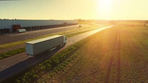 Aerial Follow Shot of White Semi Truck with Cargo Trailer Attached Moving Through Industrial Warehouse, Rural Area. Sun Shines and the Sky Are Blue. Shot on Phantom 4K UHD Camera.