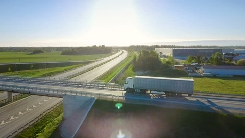  Shot on RED EPIC-W 8K Helium Cinema Camera.Aerial View of White Semi Truck with Cargo Trailer Passing Highway Overpass/ Bridge. Eighteen Wheeler is New, Loading Warehouses are Seen in the Background.