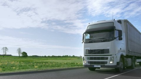 Frontal View of Speeding White Semi Truck with Cargo Trailer Drives on the Highway with Fields and Industrial Warehouses in the Background. Shot on RED EPIC-W 8K Helium Cinema Camera.