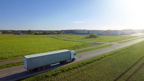 Aerial View of White Semi Truck with Cargo Trailer Moving on the Highway. In the Background Warehouses and Rural Area, Sun Shines. Shot on Phantom 4K UHD Camera.