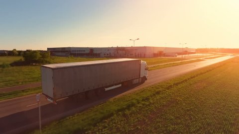Aerial View of White Semi Truck with Cargo Trailer Moving on the Highway. In the Background Warehouses and Industrial Loading Buildings are Seen. Sunset. Shot on Phantom 4K UHD Camera.