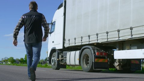 Truck Driver Crosses the Road in the Rural Area and Gets into His White Semi Truck with Cargo Trailer Attached. Sun Shines and Highway is Empty. Shot on RED EPIC-W 8K Helium Cinema Camera.