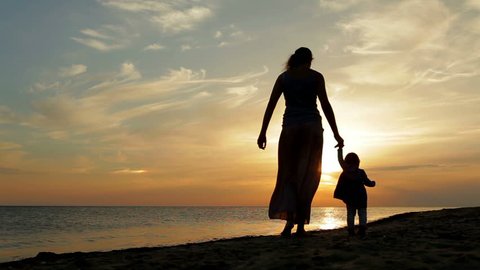Mother with baby walking on sea coast. Silhouettes sunset.