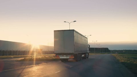 Follow-up Shot of a Semi-Truck with Cargo Trailer Moving on a Highway. White Truck Drives Through Industrial Warehouse Area on an Empty Road with Sun Shining in the Background. 4K UHD.