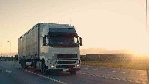 Follow-up Shot of a Semi-Truck with Cargo Trailer Moving on a Highway. White Truck Drives Through Industrial Warehouse Area in Early Hours of the Morning. Shot on RED EPIC-W 8K Helium Cinema Camera.