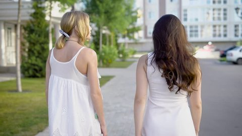 Two amazing women wearing white dresses are walking down an alley and peacfully talking about something