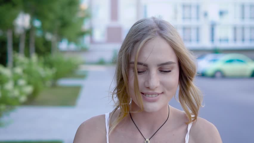 Young and unbelievably beautiful blonde woman with sky-blue eyes in white dress walks down the alley and nicely smile while combing her hair.