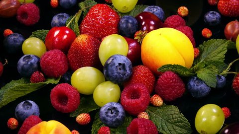 Background with mixed fresh juicy berries and fruits and mint leaves on black background in 4K. Closeup view rotation.