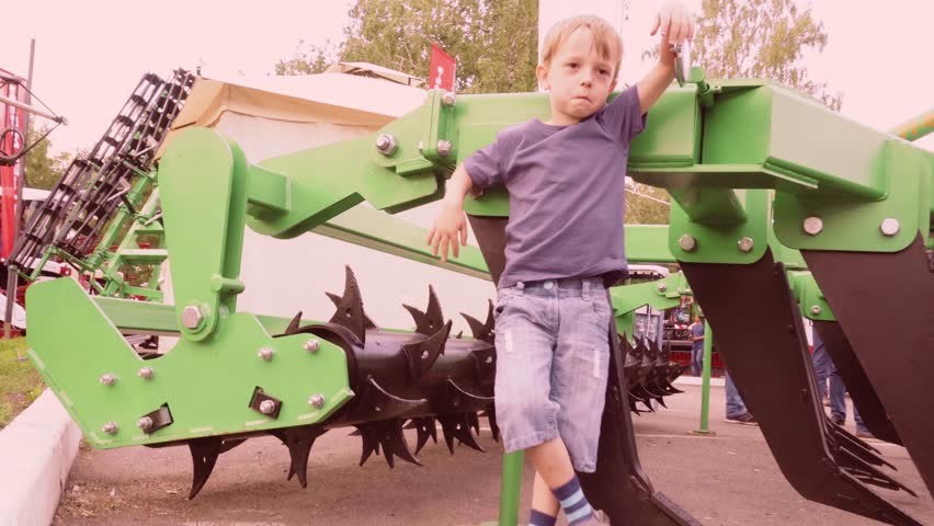 A small farmer boy stands next to agricultural machinery