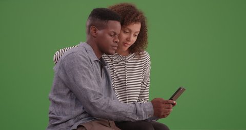 Young black couple sit on a bench looking at their smart phone on green screen. On green screen to be keyed or composited.
