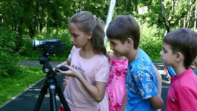 Children with video camera on tripod make movies about nature of green park. Babies outdoors in summer are creative work of cinema. Beautiful footage. Interesting to look at world in childhood.