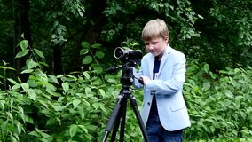 Young boy with video camera on tripod shoots film about nature slow motion. Kids outdoors in summer creative work of cinema. Beautiful footage for family. Interesting to look at world in childhood.
