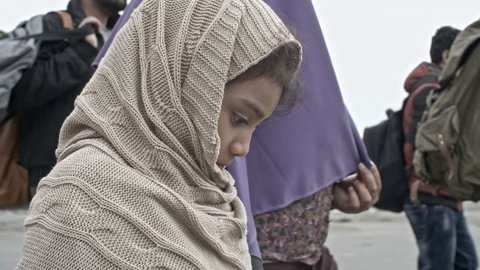 Tracking with tilt up of little Arab girl in warm poncho holding brown plush bear and walking hand in hand with her Muslim mother in purple niqab and group of refugee men
