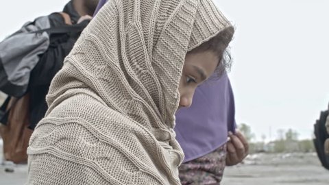 Tracking with tilt down of little Arab girl in warm poncho carrying old plush bear and holding hand of Muslim mother in niqab while walking with group of refugee men