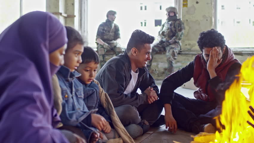 PAN of tired Arab male refugee and Muslim woman with little children wearing purple niqab warming their hands by fire in abandoned building; soldiers in uniforms chatting in background