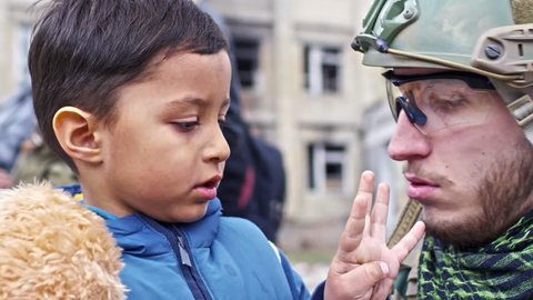Close up of Caucasian soldier in helmet and safety glasses chatting with little Syrian refugee boy with dirty face holding plush toy