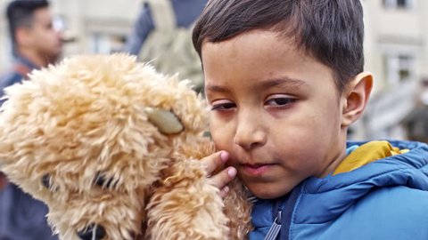 PAN with close up of sad Arab refugee boy with dirt on his face wearing puffy vest and holding plush bear