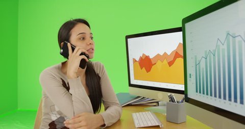 A young businesswoman talks on her mobile phone referencing the graphs on green screen. On green screen to be keyed or composited.