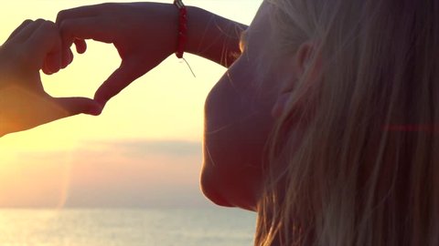 Beauty girl making heart with her hands over sea background. Happy child. Silhouette hand in heart shape with sunset inside. Slow motion 4K UHD video