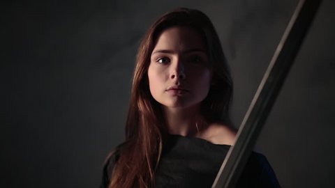 A beautiful girl with fair hair in a dark room holds a sword