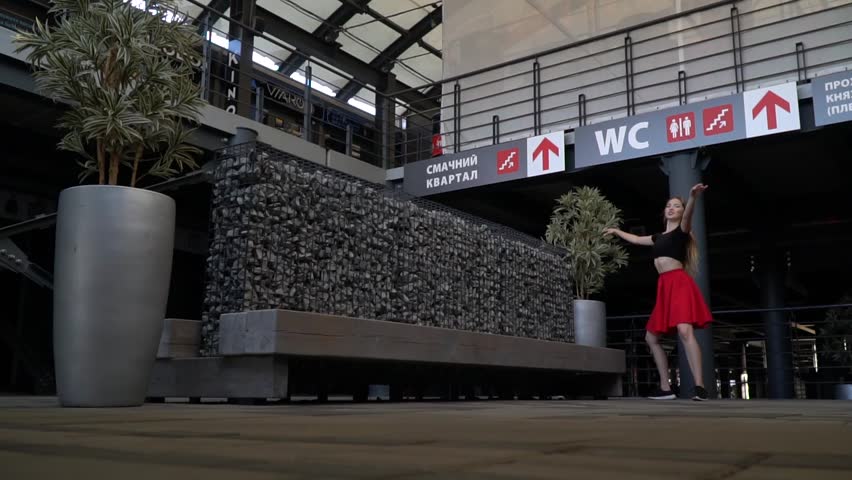 Blonde with long hair in a red skirt and a black topic is spinning in the dance in the middle of a shopping center, slow motion