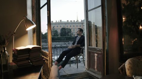 Handsome man steps out onto balcony with a book. Young man reading a book on balcony in summer evening. Attractive young man reading a book on balcony overlooking city river
