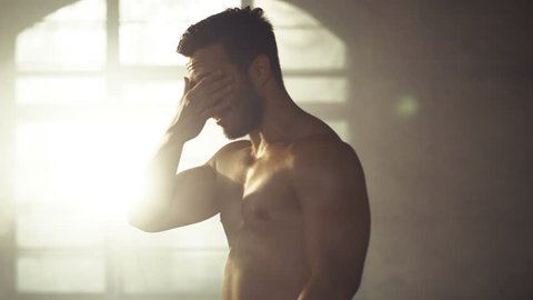 Handsome Shirtless Man with Naked Muscular Torso with Visible Six Pack Stands Resting after Bodybuilding Exercise, He Wipes Sweat from His Forehead. He's in the Middle of Abandoned Factory Building.