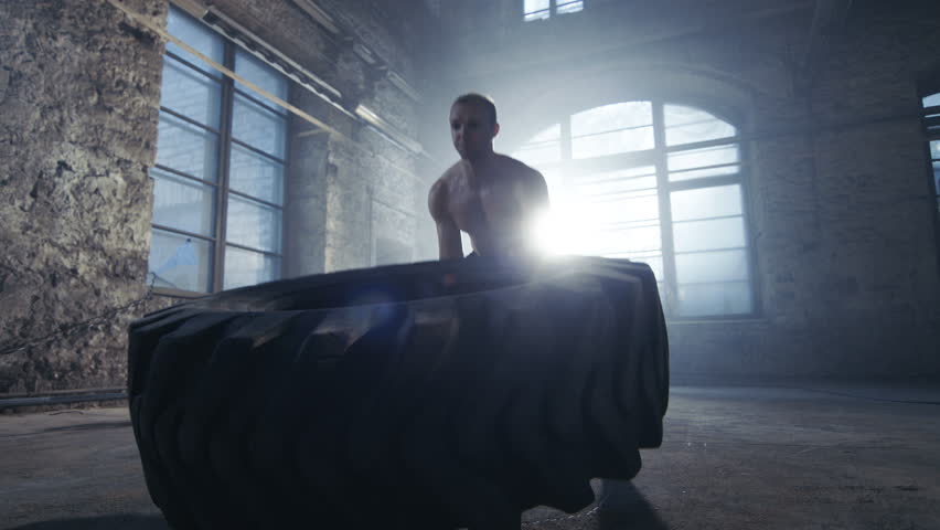 Strong Muscular Man Lifts/ Flips Tire as Part of His Bodybuilding/ Cross Fitness Training. He's Covered in Sweat and Works out in a Abandoned Factory Remodeled into Gym. Shot on RED EPIC-W 8K  Camera.