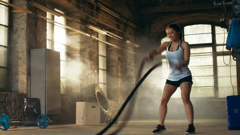 Athletic Female Actively in a Gym Exercises with Battle Ropes During Her Cross Fitness Workout/ High-Intensity Interval Training. She's Muscular and Sweaty, Gym is in Deserted Factory.