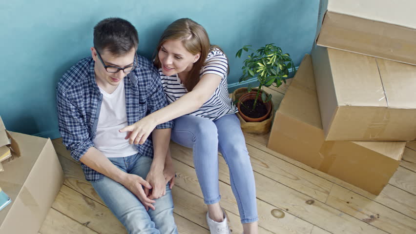 Top view of beautiful young woman and her boyfriend in glasses sitting on wooden floor of their new flat and discussing it