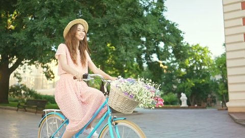 Young laughing lady in hat and pink dress riding a bicycle with flowers and smiling in the morning