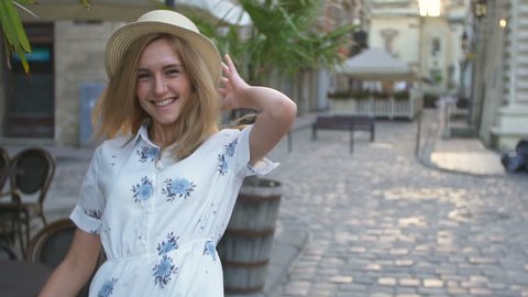Attractive girl in hat goes down the street in a city, sun is shining, than turns to camera and smiles