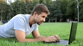 A man talking on skype in the park. A guy with a laptop lies on the grass.