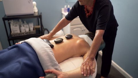 Masseuse or massage therapist uses heated stones on mature, beautiful woman’s back and hands. Steadicam