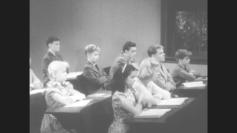 1950s: Students at desks takes notes in sync. Boy stands at desk. Woman grimaces. Woman and boy speak, boy hands woman book.