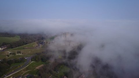 Aerial drone footage flying forwards over low lying clouds which are rolling in on top of a medieval castle in England.