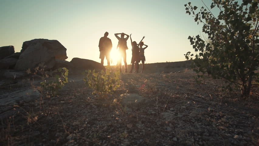 Back view of four people with backpacks posing with hands up on coastline in back lit. Slow motion