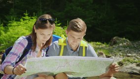 A young couple of tourists are studying together a map. They sit in a picturesque place near a mountain river. 4K video