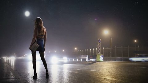 The prostitute is standing on the side of the night road where the lanterns are shining and waiting for the client. Passing cars and lights