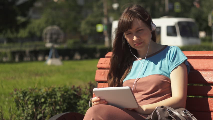 Young brunette woman listening to music with headphones sitting on a bench outside on a summer day near the road with a busy traffic